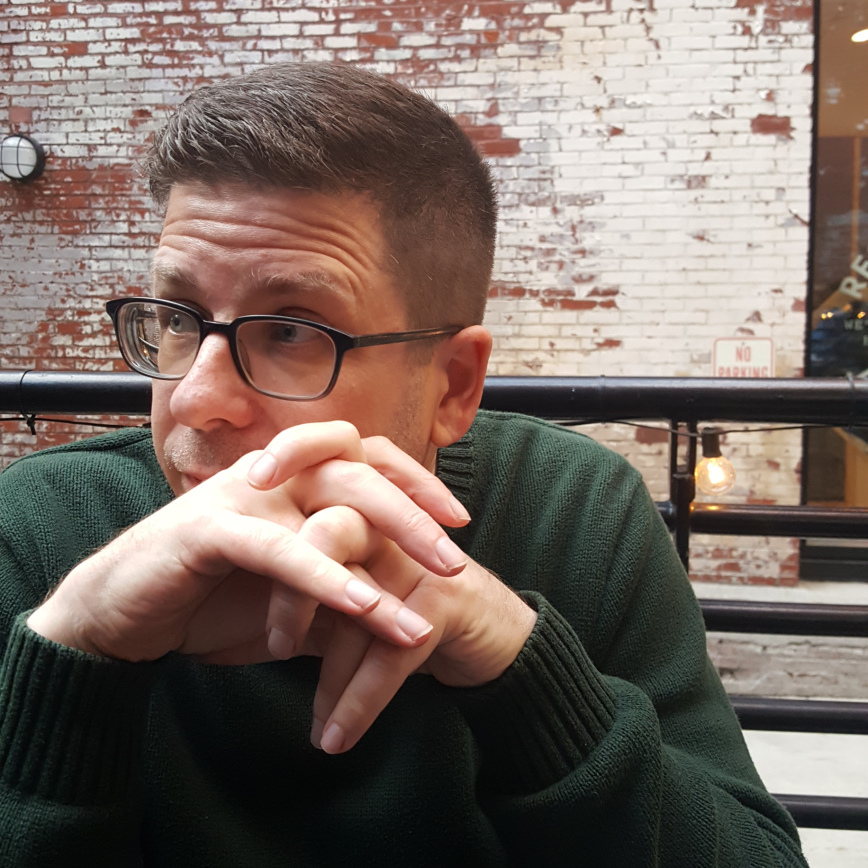 Image of Mike Caulfield at a table with his hands crossed looking his right contemplatively. 